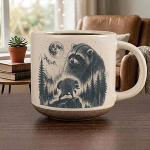 May include: A cream-colored ceramic mug featuring a dark gray illustration of a raccoon, a smaller raccoon figure on a rock, a forest of pine trees, and a full moon. The mug has a smooth handle and a slightly darker, textured base.