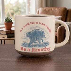 May include: A cream-colored ceramic mug with a handle, featuring a blue illustration of a raccoon in grass. The text reads in red, "in a world full of trash pandas Be a Jimothy." The base of the mug has a textured, darker brown finish.