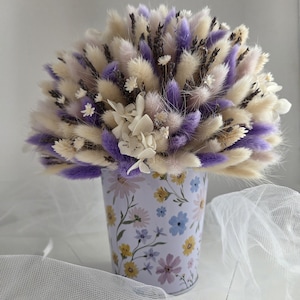 Könnte beinhalten: Ein Strauß aus Trockenblumen mit flauschigen cremefarbenen und violetten Hasenschwanzgräsern, Lavendelzweigen und kleinen weißen Blüten. Das Arrangement steht in einem zylindrischen Metallbehälter mit einem bunten Blumenmuster.