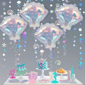 May include: A party display featuring four iridescent seashell-shaped foil balloons suspended above a table. The table holds a cake with a scale pattern, cupcakes with mermaid tail toppers, sea-themed decorations, and various party supplies.
