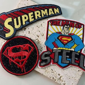 Può includere: Tre toppe ricamate a tema Superman. Una toppa mostra la parola SUPERMAN in giallo su sfondo rosso. Un'altra è una toppa circolare nera con uno scudo S rosso. La terza mostra un Superman dei cartoni animati con il testo THE MAN OF STEEL.