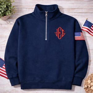 May include: A navy blue quarter-zip pullover featuring a stand-up collar with a silver zipper. The chest displays an embroidered red monogram with the letters J, E, and E in a diamond shape. A small American flag patch is on the right sleeve.