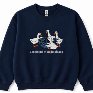 Può includere: Una felpa girocollo blu navy con un ricamo raffigurante quattro oche bianche riunite attorno a un piccolo computer portatile. Sotto le oche, il testo "a moment of code please" è ricamato in bianco. Presenta maniche lunghe e polsini a costine.