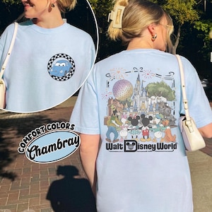 Puede incluir: Una camiseta azul claro con un peque&ntilde;o gr&aacute;fico circular en la parte delantera que muestra un coche azul dentro de un borde a cuadros. La parte trasera muestra una gran ilustraci&oacute;n de personajes Disney frente a un castillo y el texto Walt Disney World.