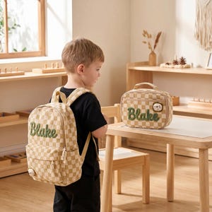 Puede incluir: Una mochila de pana a cuadros beige y blancos y una bolsa de almuerzo rectangular a juego. Ambos artículos llevan el nombre Blake bordado en letra verde, con un pequeño león de peluche en la bolsa de almuerzo y un pequeño dije decorativo.