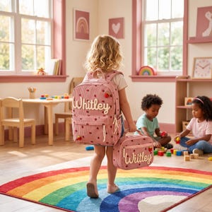 Puede incluir: Una mochila y una bolsa de almuerzo a juego de pana rosa polvoriento con un patrón de fresas repetido. Ambos artículos están personalizados con el nombre Whitley en letras cursivas blancas e incluyen tiradores de cremallera con forma de fresa.