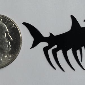 May include: A black silhouette of a hammerhead shark with multiple thin, pointed legs extending from its underside, positioned next to a United States quarter coin for scale to show its size relative to the 0.955 inch diameter currency.