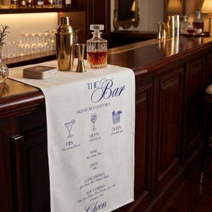 Könnte beinhalten: Ein weißer Tischläufer aus Stoff mit blauem Aufdruck von Text und Cocktailglas-Illustrationen. Der Text lautet The Bar, Signature Cocktails, His, Hers, Ours, Wine, Beer, Long Drinks, Soft Drinks und Cheers. Er liegt auf einer hölzernen Bar.