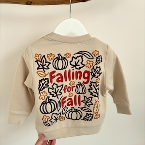 May include: A beige long-sleeve sweatshirt featuring a graphic print of pumpkins, leaves, and flowers in brown, orange, and red. The text "Falling for Fall" is printed in red in the centre of the design, surrounded by the autumn-themed illustrations.