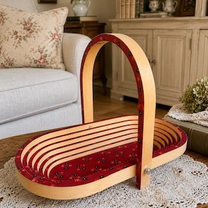 May include: An oval wooden basket with a collapsible design, featuring a light natural wood exterior and a deep red interior decorated with a small black and red floral pattern. The basket has a single arched handle attached with metal hardware.