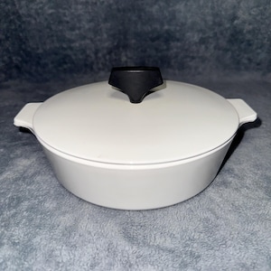 May include: A white round casserole dish with a matching lid and two side handles. The lid features a black, triangular-shaped knob handle centered on top. The dish has a smooth, solid white finish and a low-profile, wide-rimmed design.