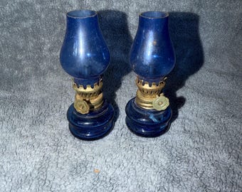 Könnte beinhalten: Ein Paar passende Öllampen mit tiefblauen Glassockeln und passenden blauen Glaszylindern. Jede Lampe verfügt über eine goldfarbene Metallbrennereinheit mit einem runden Einstellknopf an der Seite des zentralen Metallkragens.