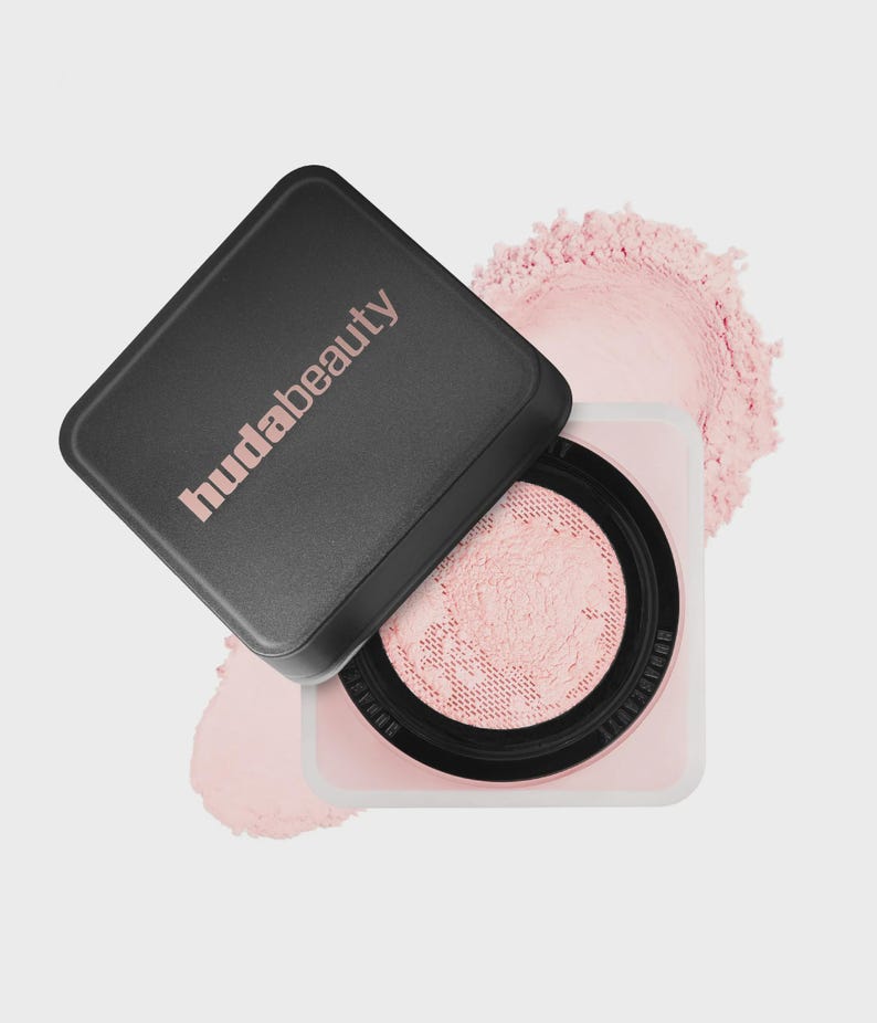 May include: A square cosmetic container with a black lid featuring the text hudabeauty in light pink. The base is light pink with a black circular mesh sifter containing loose light pink powder, with additional powder spilled around the base.
