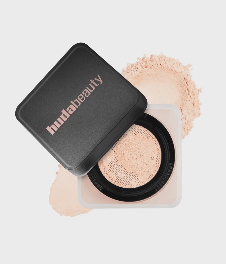 May include: A square cosmetic container with a black lid featuring the text hudabeauty in light pink. The base is clear with a black inner rim and a mesh sifter containing loose, light beige setting powder, with more powder visible behind it.