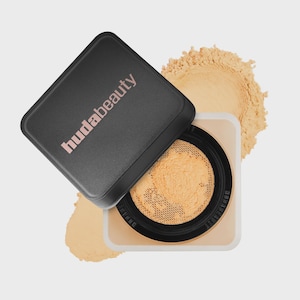 May include: A square Huda Beauty loose setting powder container with a black lid featuring the brand name in light pink text. The lid is tilted, revealing a beige powder inside a mesh sifter, with a pile of loose beige powder visible behind it.