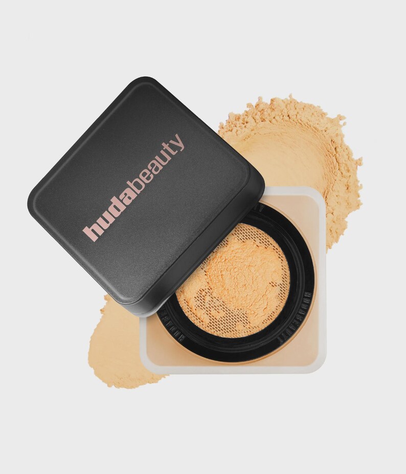 May include: A square Huda Beauty loose setting powder container with a black lid featuring the brand name in light pink text. The lid is tilted, revealing a beige powder inside a mesh sifter, with a pile of loose beige powder visible behind it.