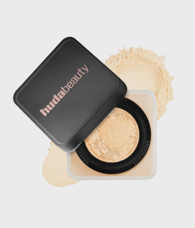 May include: A square cosmetic container with a black lid featuring the text hudabeauty in light pink. The base is clear with a black inner rim and a mesh sifter containing loose beige setting powder, with additional powder visible behind it.