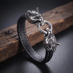 Könnte beinhalten: Ein schwarzes Lederarmband mit zwei detaillierten silberfarbenen Wolfsköpfen an den Enden. Die Wolfsköpfe sind durch einen runden silberfarbenen Metallringverschluss verbunden. Das Lederband hat sichtbare weiße Nähte und eine geflochtene Struktur.