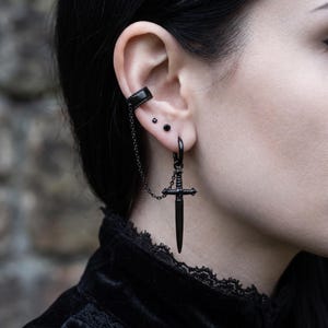 Puede incluir: Un pendiente tipo ear cuff negro conectado por una cadena fina a un aro negro con un colgante de espada. La espada tiene una empuñadura detallada y una hoja puntiaguda. Dos pequeños pendientes de botón negros se sitúan entre ambos.