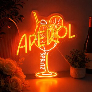 Peut inclure: Une enseigne néon orange vif en forme de verre à cocktail avec une paille et une tranche d'orange. L'enseigne affiche le texte APEROL en grandes lettres et le mot SPRITZ verticalement le long du pied du verre, brillant sur un mur sombre.