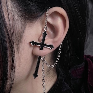 May include: A silver-toned ear cuff featuring a large cross pendant with black enamel inlay. The cross is connected to a delicate silver chain that loops from the top of the ear to the base of the cross, creating a layered, dangling effect.