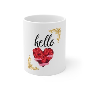 Puede incluir: Una taza de cerámica blanca con la palabra hello en fuente cursiva negra. Debajo del texto hay un arreglo de rosas rojas en forma de corazón envuelto en una cinta rosa claro. El diseño está enmarcado por dos adornos dorados en filigrana.
