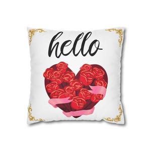 Puede incluir: Un cojín cuadrado blanco con la palabra hello en letra cursiva negra en la parte superior. Debajo del texto hay un arreglo en forma de corazón de rosas rojas envueltas en una cinta rosa claro. Las esquinas tienen detalles dorados.