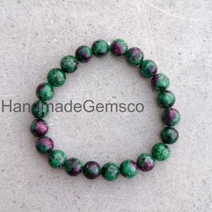 May include: A circular beaded bracelet composed of round, polished gemstones featuring a mottled pattern of deep forest green and vibrant magenta-pink. The beads are strung together on an elastic cord. The text HandmadeGemsco is visible.