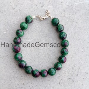 May include: A beaded bracelet featuring round, polished gemstones with a mottled pattern of deep forest green and vibrant magenta-pink. The beads are strung together and secured with a silver-toned metal lobster claw clasp and a short chain extender.