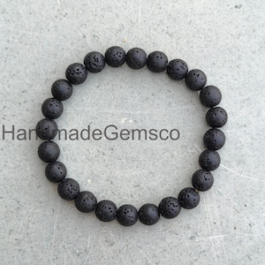 Puede incluir: Una pulsera circular compuesta por numerosas cuentas pequeñas y redondas de piedra de lava negra con una superficie porosa y texturizada. Las cuentas están ensartadas en un bucle continuo. El texto HandmadeGemsco es visible en el centro.