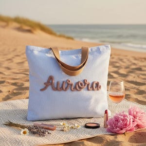Puede incluir: Un bolso tote de seersucker en azul claro y blanco con asas de color tostado. La palabra Aurora está bordada en una letra cursiva rosa polvoriento en relieve en el centro del bolso. El bolso es rectangular con base plana y dos asas.