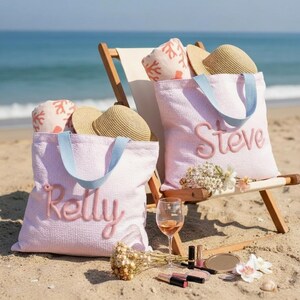 Puede incluir: Dos bolsos tote de seersucker rosa claro con asas azul claro, cada uno con los nombres bordados 'Kelly' y 'Steve'. Cada bolso contiene un sombrero de paja y una toalla con estampado de coral. Una copa de vino y maquillaje están cerca.