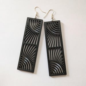 Könnte beinhalten: Ein Paar schwarze, rechteckige Ohrhänger mit einem lasergeschnittenen Design aus zwei halbkreisförmigen Sonnenstrahlenmustern mit strahlenden Linien und konzentrischen Bögen. Die Ohrringe sind an silberfarbenen Metall-Ohrhaken befestigt.