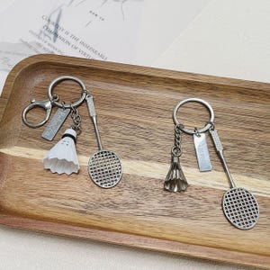 Peut inclure: Deux porte-clés en métal argenté avec des breloques sur le thème du badminton. Chaque porte-clés comprend une raquette de badminton miniature, un volant et une plaque rectangulaire gravée avec les prénoms Rebecca et Anne.