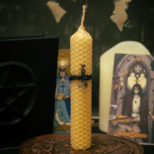 May include: A cylindrical beeswax candle with a textured honeycomb pattern, featuring a natural jute wick at the top and a thin black string tied around the center. The candle stands upright on a circular wooden base with carved floral details.