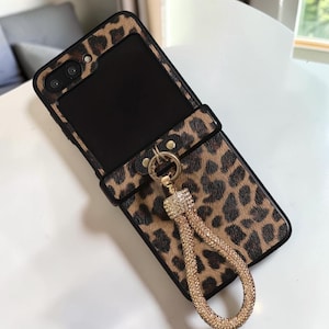 May include: A rectangular phone case featuring a brown and black leopard print pattern with a black border. A matching leopard print strap attachment is secured with a gold-toned metal ring and a loop covered in small, clear, sparkling rhinestones.