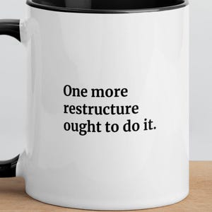 Op de afbeelding: Een witte keramische mok met een zwarte binnenkant en een zwart handvat. Op de voorzijde van de mok staat de zwarte tekst: One more restructure ought to do it. De mok heeft een cilindrische vorm en een gladde, glanzende afwerking aan de buitenkant.