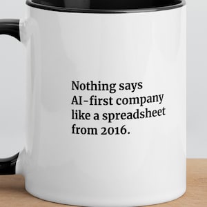 Könnte beinhalten: Ein weißer Keramikbecher mit schwarzem Innenraum und Henkel. Auf der Vorderseite steht in schwarzer Schrift: Nothing says AI-first company like a spreadsheet from 2016. Der Becher hat eine glatte, glänzende Oberfläche und eine zylindrische Form.