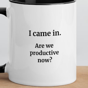 Peut inclure: Un mug en céramique blanche avec une anse et un bord noirs. L'avant du mug présente un texte imprimé en noir qui dit : I came in. Are we productive now ? Le motif est centré sur la surface cylindrique lisse de cet objet de vaisselle.