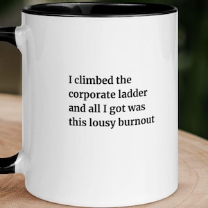 Puede incluir: Una taza de cerámica blanca con interior y asa de color negro. La parte frontal de la taza presenta un texto impreso en negro que dice: I climbed the corporate ladder and all I got was this lousy burnout. La taza tiene un acabado liso.