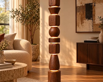Può includere: Una lampada da terra alta caratterizzata da una pila verticale di forme geometriche in legno marrone scuro, inclusi cubi, sfere e cilindri, con una luce calda che emana dagli spazi orizzontali tra ogni segmento in legno.