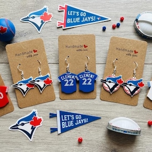 May include: Four pairs of Toronto Blue Jays themed earrings on brown cardboard displays. Designs include red Kirk 30 jerseys, blue jay bird heads, blue Clement 22 jerseys, circular team logos, and white Vladdy 27 jerseys. Includes pennant stickers and beads.