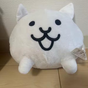 May include: A white plush toy shaped like a round cat with pointed ears. The face features two small black circular eyes and a wide, black embroidered mouth with a tongue. Two small white paws extend from the bottom of the rounded body.