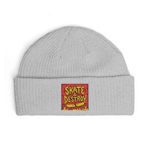 May include: A light gray knit beanie featuring a square embroidered patch on the front cuff. The patch displays the text "SKATE & DESTROY" in yellow letters over a red background, with a yellow broken skateboard graphic and orange flames below.