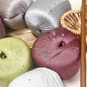 May include: Several round balls of yarn in shades of mauve, light green, silver, and white, each featuring a subtle sheen and small, scattered reflective sequins. A single wooden knitting needle is positioned vertically along the right side.