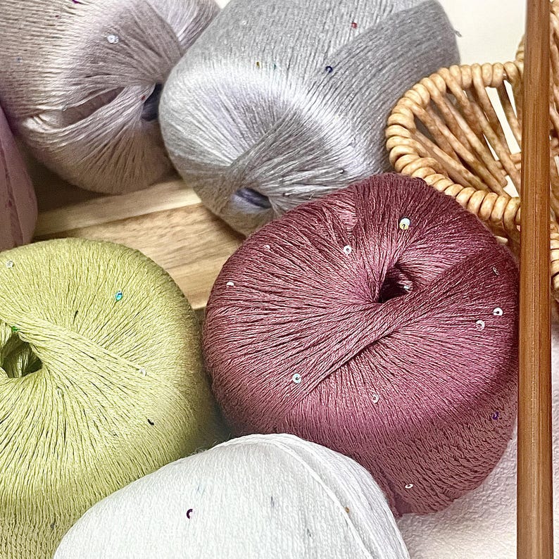 May include: Several round balls of yarn in shades of mauve, light green, silver, and white, each featuring a subtle sheen and small, scattered reflective sequins. A single wooden knitting needle is positioned vertically along the right side.