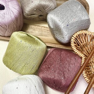 May include: Six balls of yarn in various colors including light purple, beige, silver, olive green, dusty rose, and white. Each ball of yarn features a subtle sheen and is embellished with small, scattered sequins throughout the fiber strands.