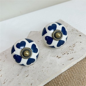 Peut inclure: Deux boutons de placard ronds en céramique avec une base blanche et un motif de cœurs bleu foncé répété. Chaque bouton possède une fixation centrale circulaire en métal couleur laiton vieilli avec un petit dôme en relief.