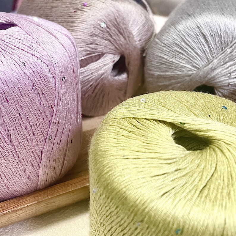 May include: Four round balls of yarn in pastel shades of light purple, soft pink, pale green, and light gray. Each ball of yarn features a smooth, slightly shiny texture with small, scattered reflective sequins embedded throughout the fibers.