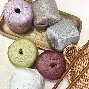 May include: Six balls of yarn in lavender, beige, silver, light green, dark pink, and white, each featuring small embedded sequins. The yarn balls are arranged on a wooden tray alongside a woven wicker basket and two long wooden knitting needles.